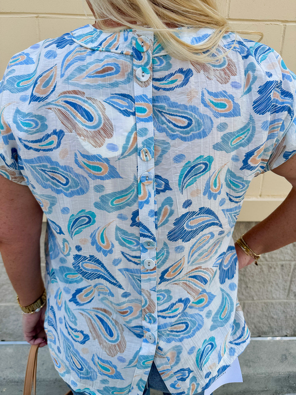 Blue paisley print cap sleeve blouse with round neckline and back button detail, styled for a soft and polished look.
