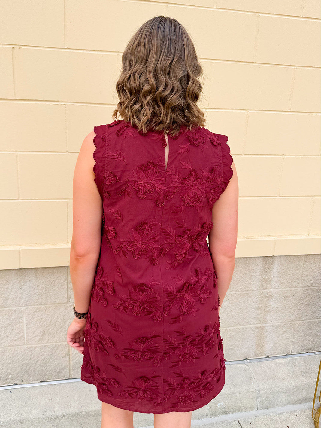 Sleeveless mini dress in rich garnet with 3D floral appliqué, scalloped embroidered hem, and side pockets. Elegant, textured, and perfect for fall or Gameday.