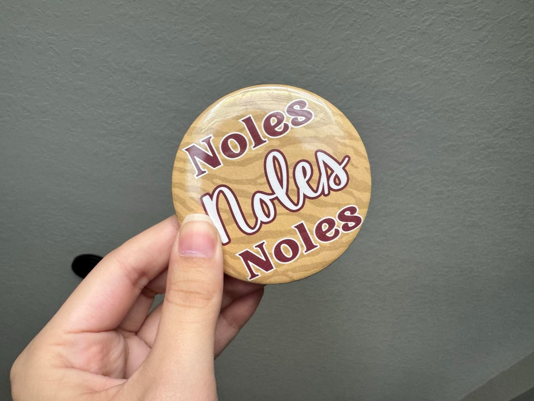 Round button with garnet “Noles, Noles, Noles” lettering on a subtle gold animal print background, styled as a gameday bag accessory.