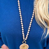 Tan beaded necklace with gold accents and a layered hammered gold and tan pendant, styled for versatile wear.