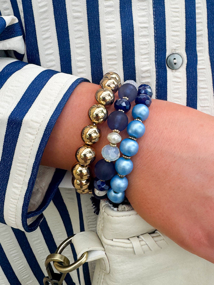 Layered charm bracelet stack featuring blue and gold glass beads, gold accents, and a tassel pendant for a stylish, boho-inspired look.