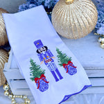 White tea towel with blue and white nutcracker, red drum, ginger jar Christmas trees, and blue scalloped trim.