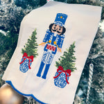 White tea towel with blue and white nutcracker, red drum, ginger jar Christmas trees, and blue scalloped trim.