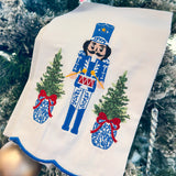 White tea towel with blue and white nutcracker, red drum, ginger jar Christmas trees, and blue scalloped trim.