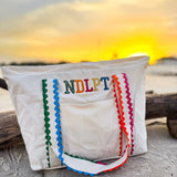 Lightweight canvas NDLPT project tote with embroidered design, colorful ric-rac shoulder straps, zip-top closure, and exterior pocket