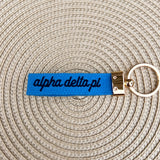 Double-sided embroidered Alpha Delta Pi sorority keychain with navy Greek letters, script sorority name, white strap, and gold hardware.