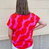 Banana and floral print top in orange and hot pink with V-neck and short dolman sleeves, displayed on hanger against neutral background