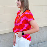 Banana and floral print top in orange and hot pink with V-neck and short dolman sleeves, displayed on hanger against neutral background