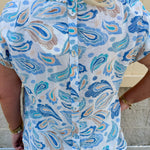 Blue paisley print cap sleeve blouse with round neckline and back button detail, styled for a soft and polished look.
