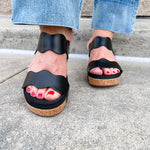 Black scalloped wedge sandals with cork sole, gold hardware accents, and dual straps—stylish and comfortable for everyday wear.