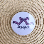 Delta Gamma button pin featuring a pink bow with cursive “Dee Gee” lettering on a white background.
