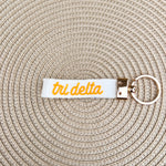 Double-sided embroidered Delta Delta Delta sorority keychain with white strap, signature color Greek letters, sorority name, and gold hardware.