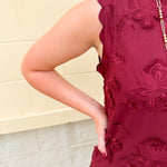 Sleeveless mini dress in rich garnet with 3D floral appliqué, scalloped embroidered hem, and side pockets. Elegant, textured, and perfect for fall or Gameday.