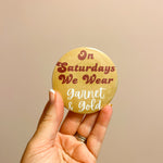 Gold button with garnet and white lettering reading “On Saturdays We Wear” styled for FSU game day.