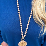 Tan beaded necklace with gold accents and a layered hammered gold and tan pendant, styled for versatile wear.