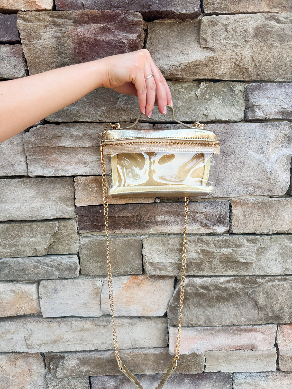 Clear stadium-approved bag with gold vegan leather accents, gold chain strap, and zipper.