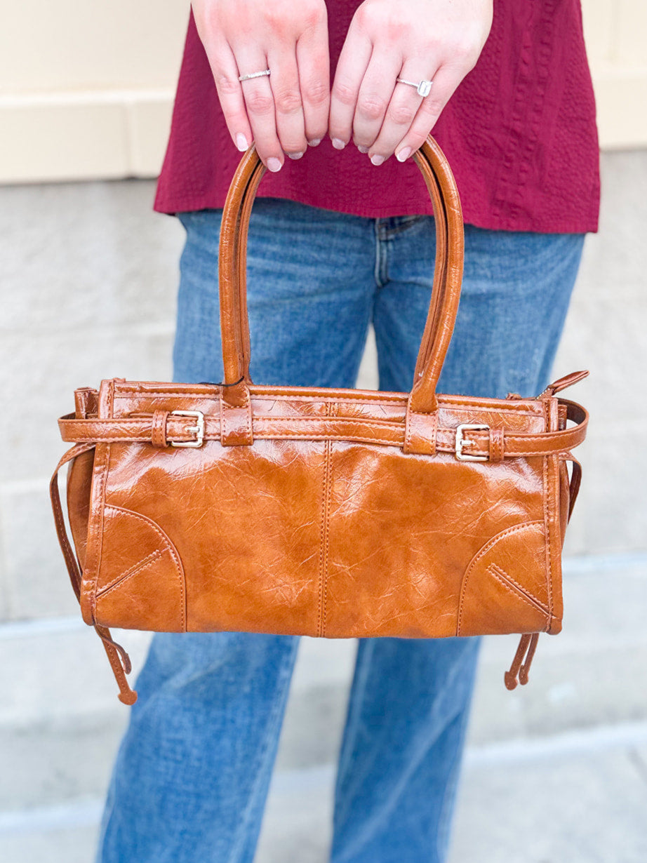 Structured caramel brown glossy faux leather tote with rounded handles and buckle accents.