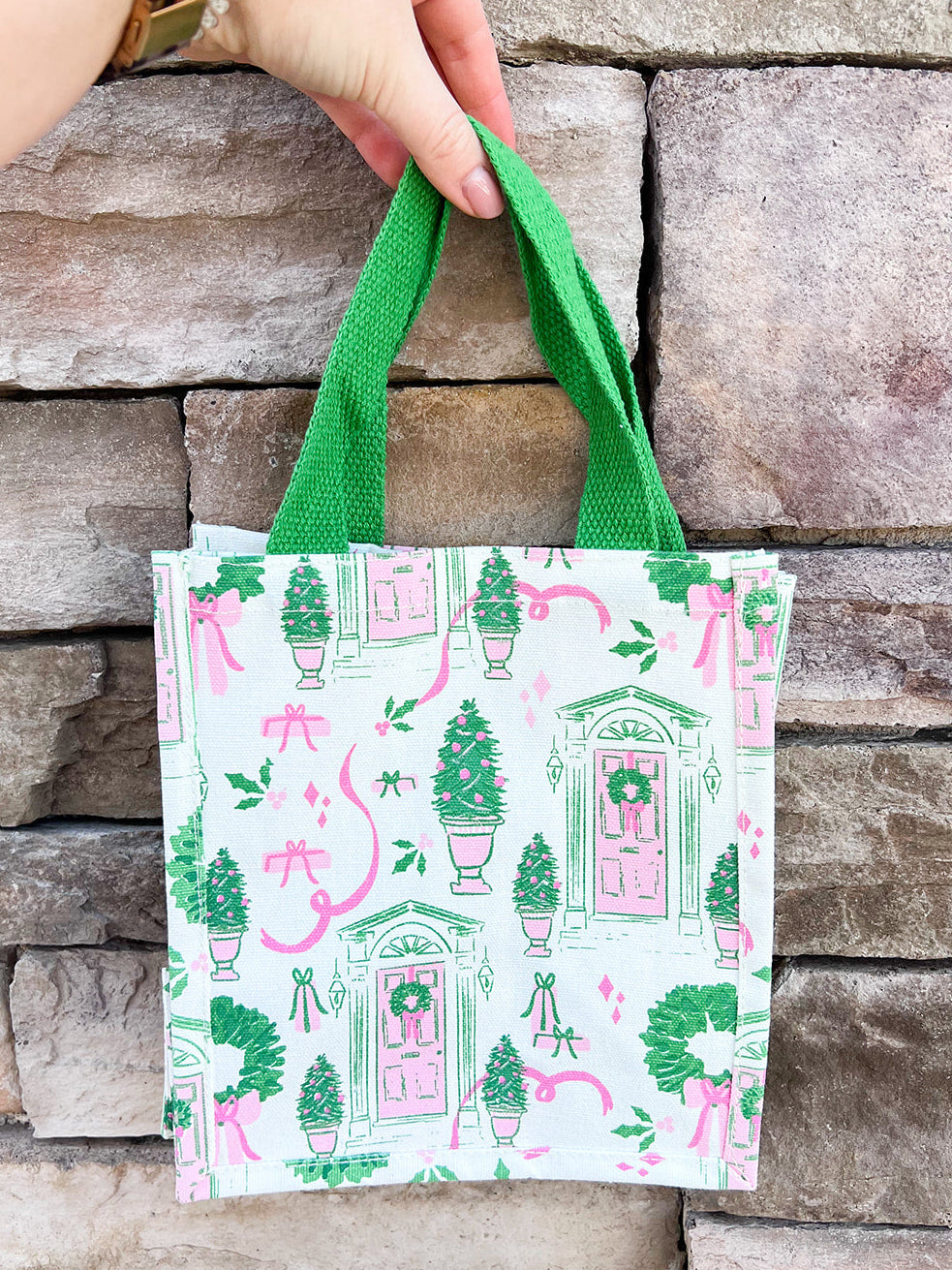 White compact tote with pink and green holiday print featuring front doors, wreaths, potted trees, and green handles.