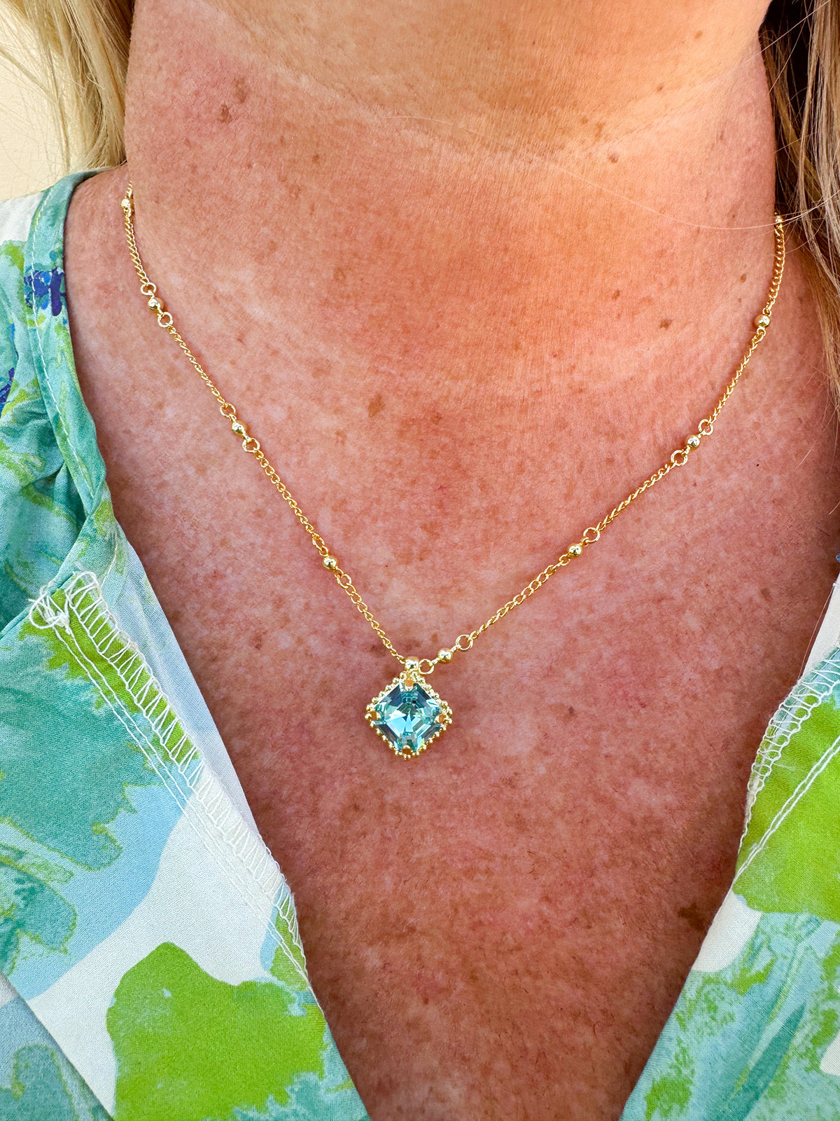 Bright gold-tone necklace featuring a square-cut aquamarine crystal with metal beaded halo on an adjustable chain.