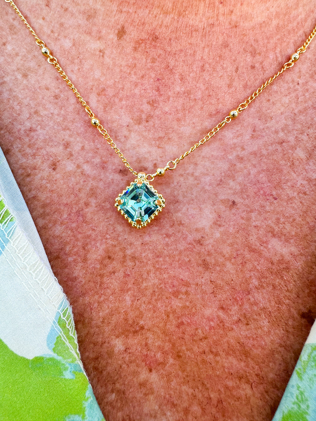 Bright gold-tone necklace featuring a square-cut aquamarine crystal with metal beaded halo on an adjustable chain.