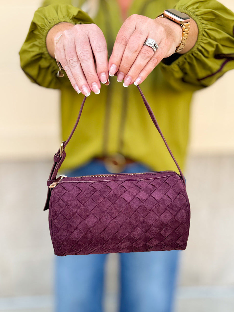 Plum-colored cylinder-shaped shoulder bag with adjustable buckle strap and zip closure.