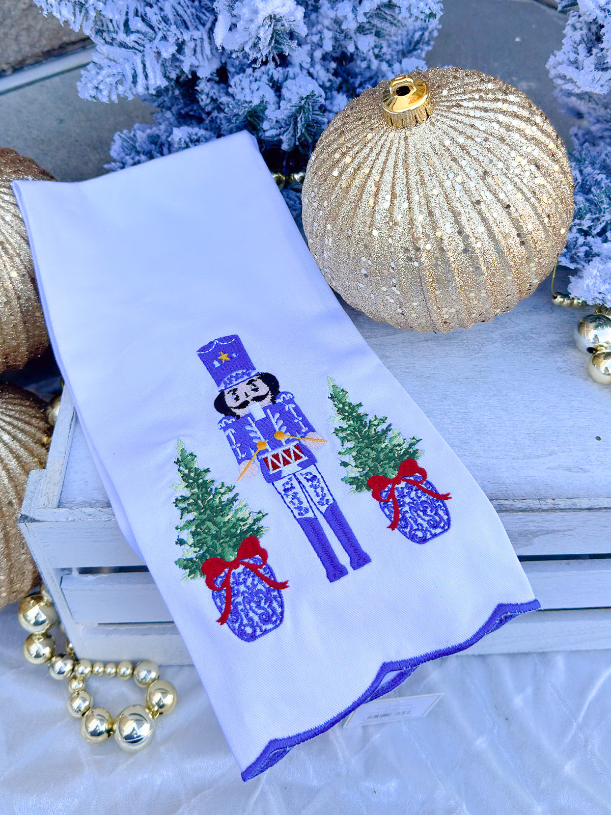 White tea towel with blue and white nutcracker, red drum, ginger jar Christmas trees, and blue scalloped trim.
