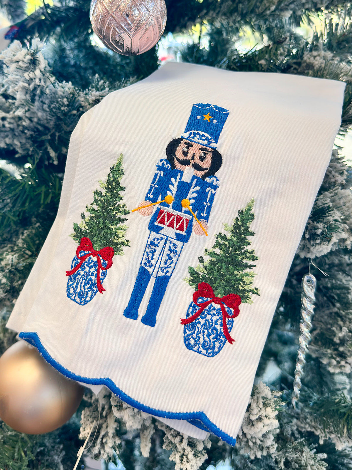 White tea towel with blue and white nutcracker, red drum, ginger jar Christmas trees, and blue scalloped trim.