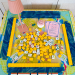 A colorful mahjong game board of the Pippa tiles on the Hapton Mat setup photographed from above, with a pink lamp, scattered yellow tiles, white tiles, blue border pieces, and instruction cards on a light wood surface.