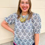Women’s ivory top with navy and light blue porcelain-inspired print, short puff sleeves, gathered neckline, and hot pink scalloped hem trim.