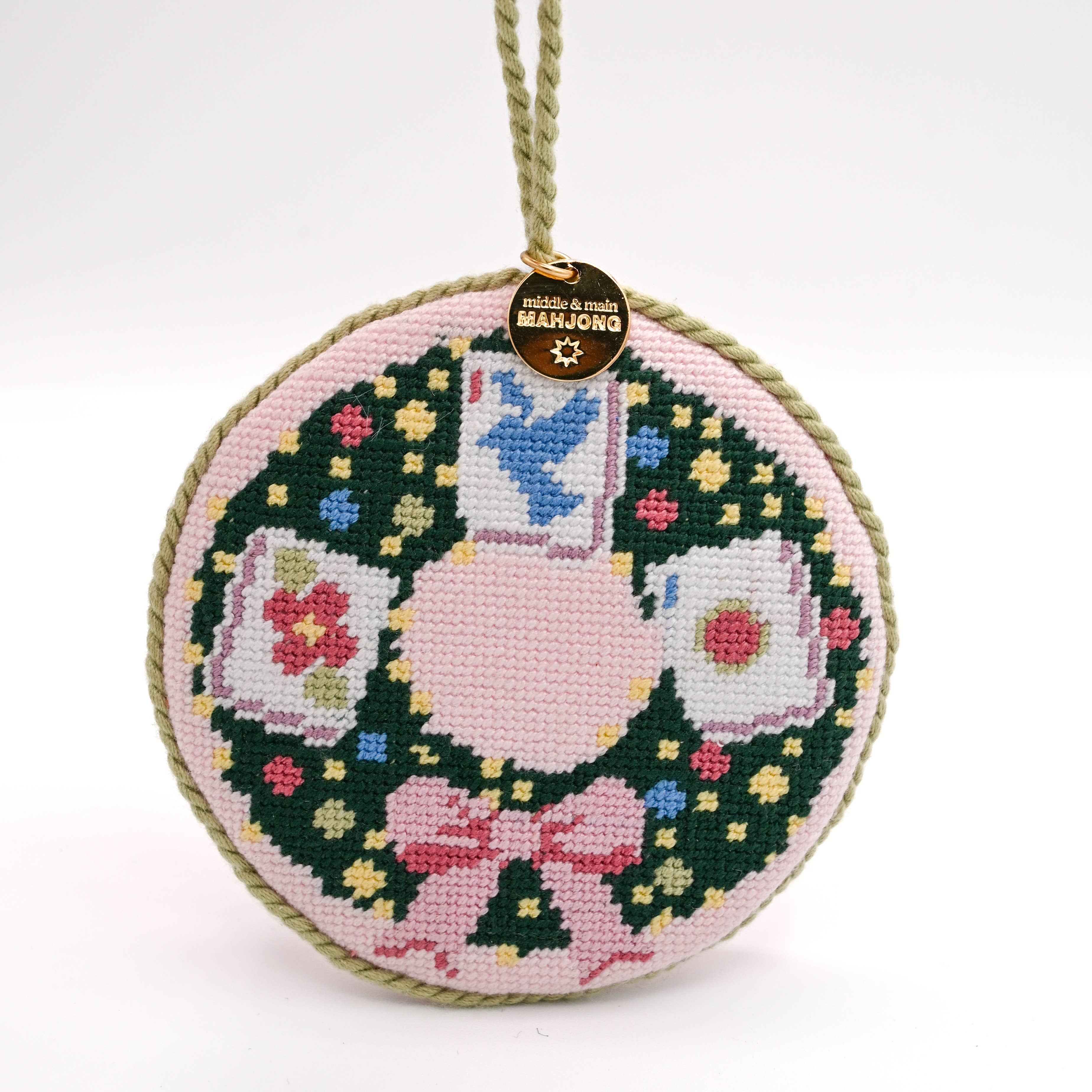 Round needlepoint mahjong ornament with wreath design, colorful mahjong tiles, bird, and pink bow.