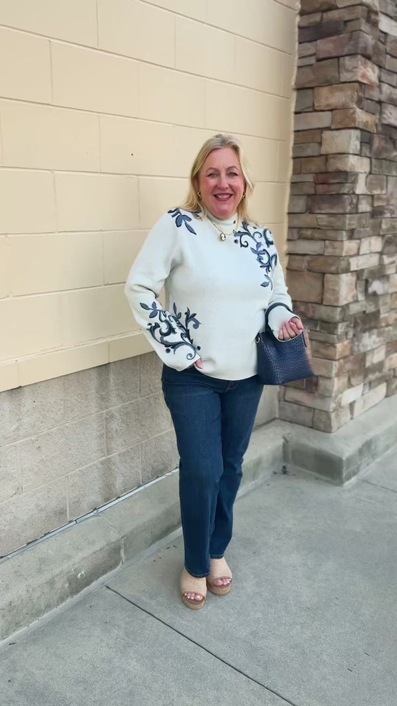 White long-sleeve mock-neck sweater with navy and light blue floral details on the shoulders and sleeves.