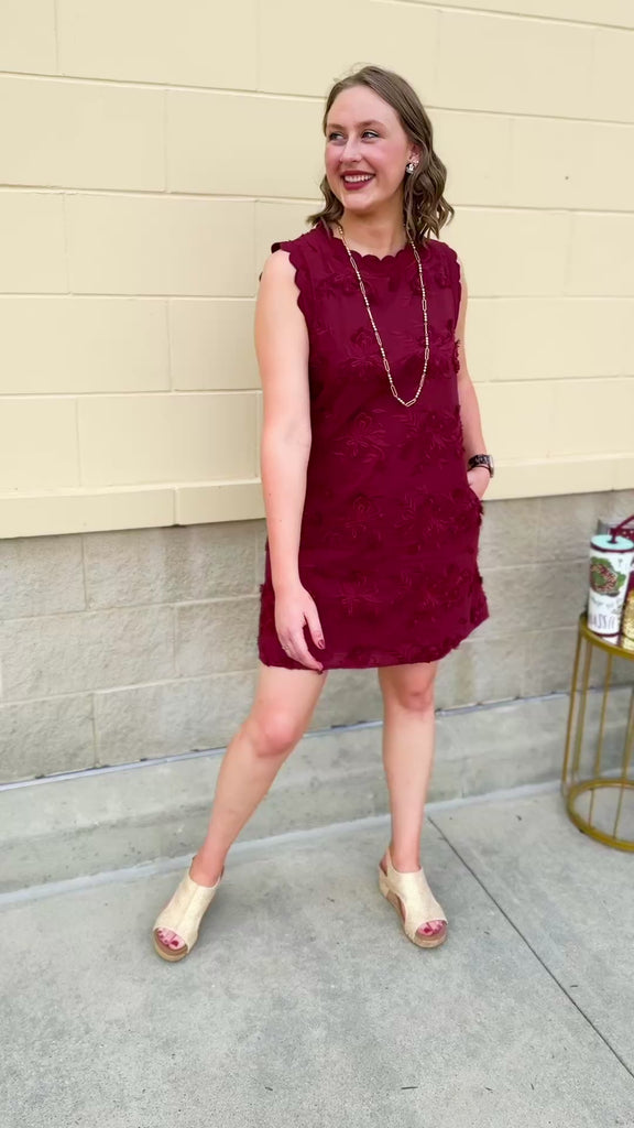 Sleeveless mini dress in rich garnet with 3D floral appliqué, scalloped embroidered hem, and side pockets. Elegant, textured, and perfect for fall or Gameday.